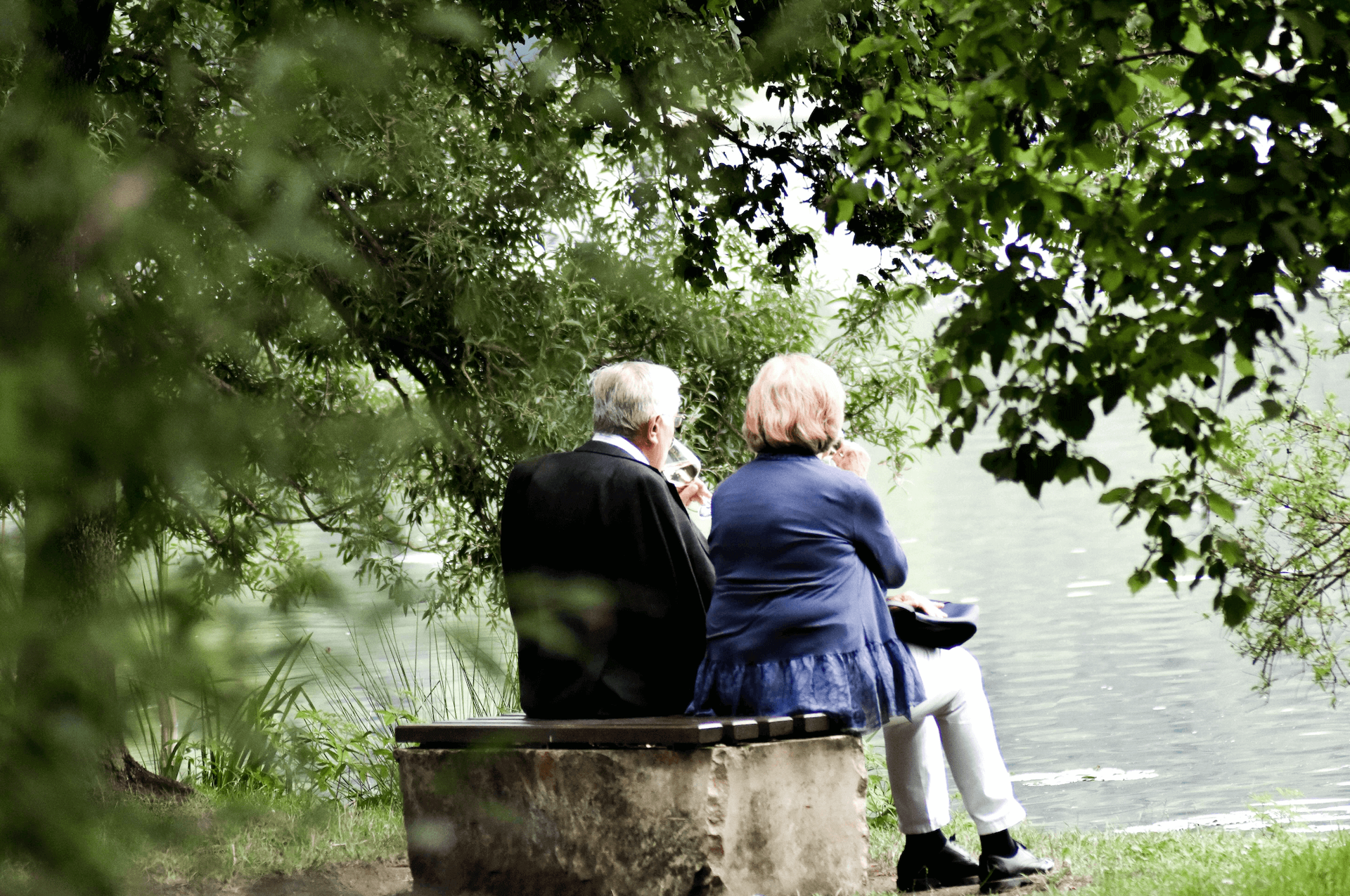 älteres Ehepaar sitzt auf einer Bank und blickt auf einen See.