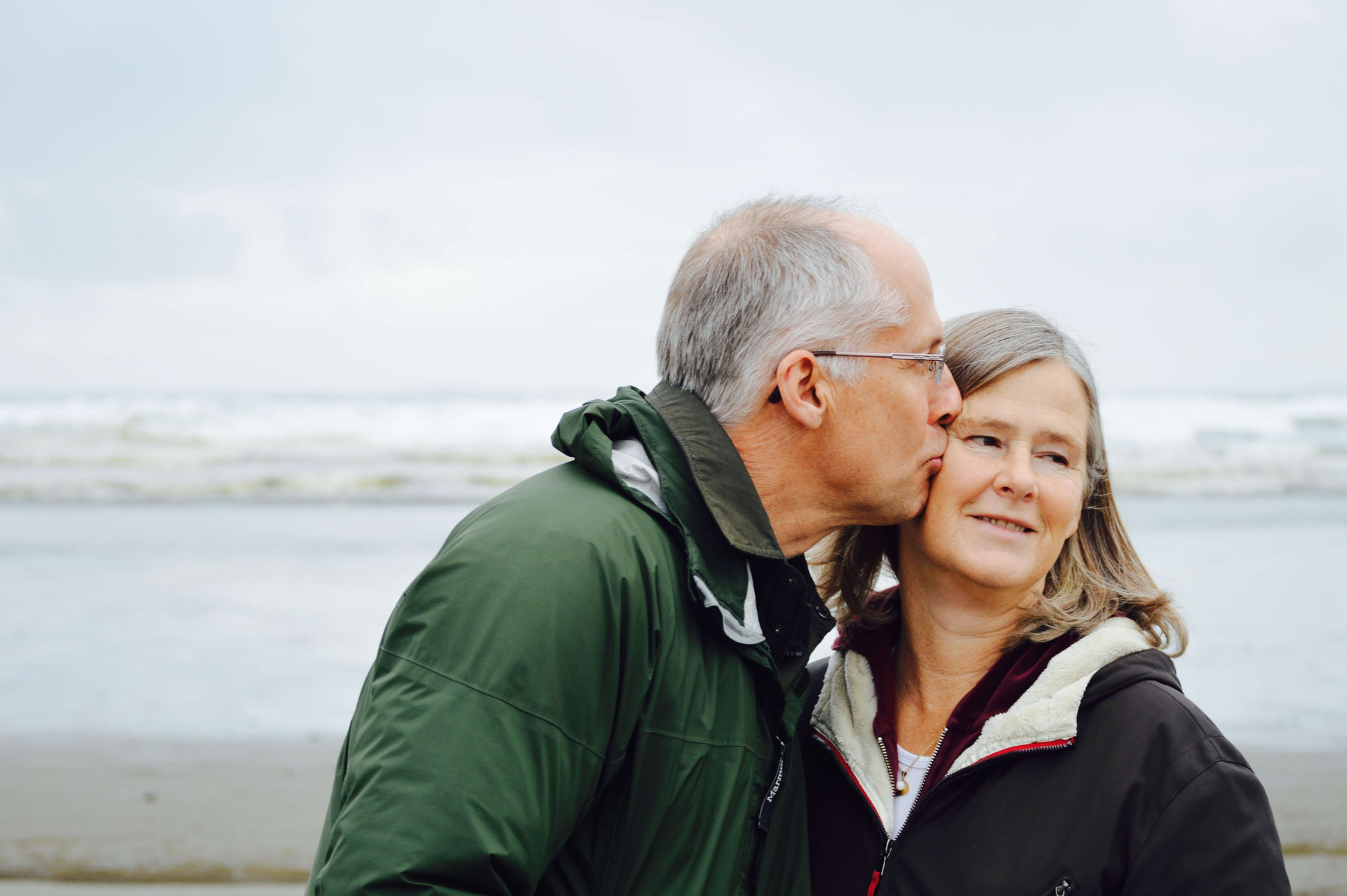 Ehemann küsst seine Ehefrau auf die Wange. Im Hintergrund raues Meer am Strand.